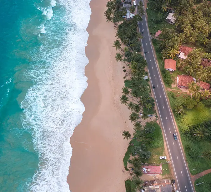 Coastal road beside the Indian Ocean in Weligama, Sri Lanka, with waves meeting sandy shores.