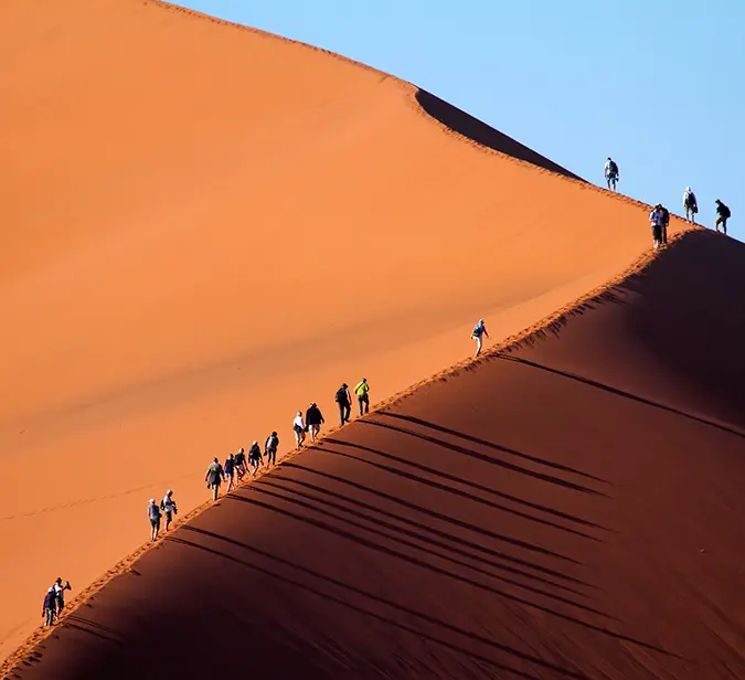 Travellers hiking up a tall sand dune in Sossusvlei at sunrise in Namib-Naukluft National Park.