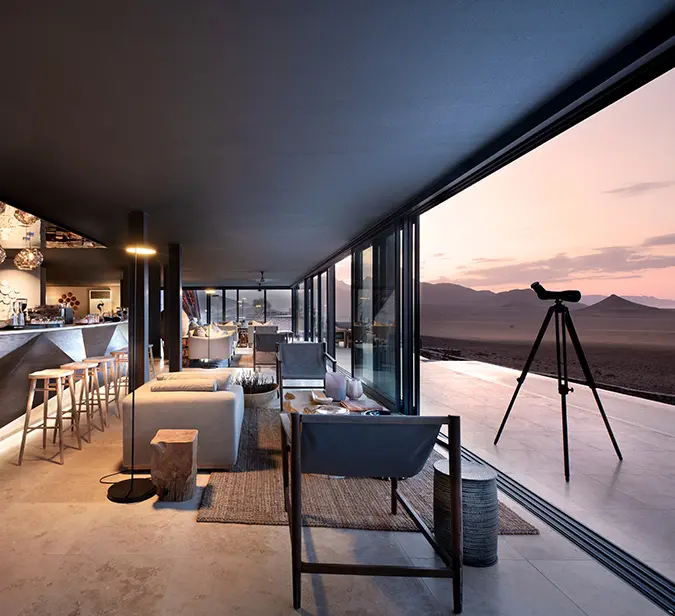 Luxury desert lodge lounge with panoramic views of Sossusvlei dunes at sunset in Namibia.