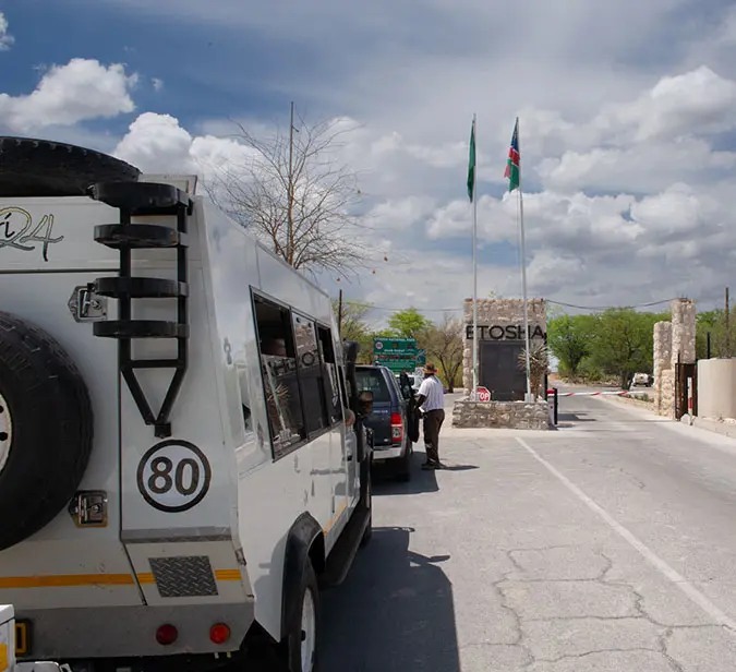 Vehicles queue at entrance gate of Etosha National Park under blue sky with Namibian flags flying.
