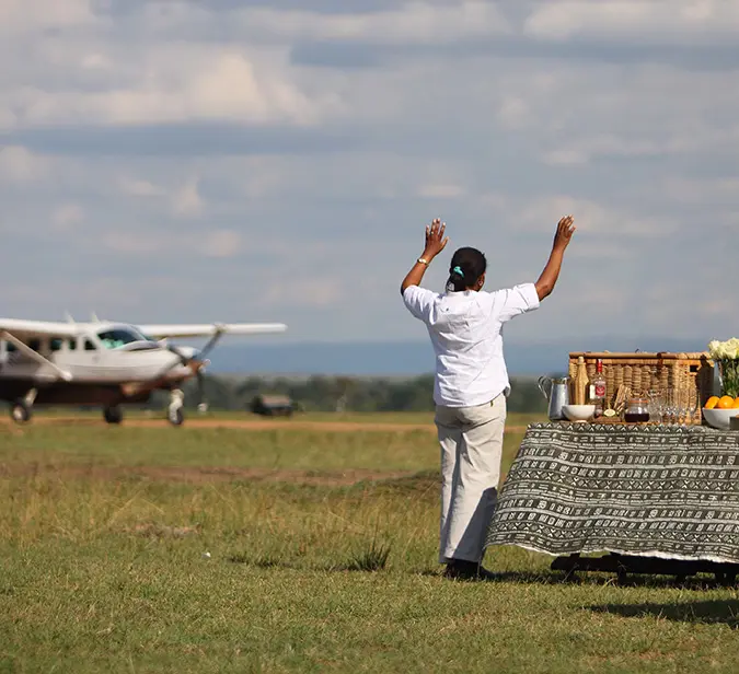 Safari staff bids goodbye beside bush airstrip with bush plane and breakfast table in view.