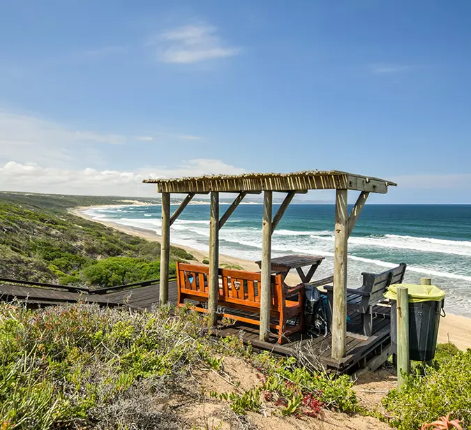 Oceanfront resting spot with wooden bench and pergola on South Africa’s coastal hiking route.