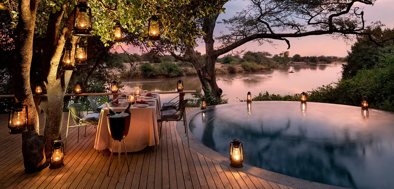 Luxury safari dinner by pool and river at sunset, surrounded by lanterns and nature.